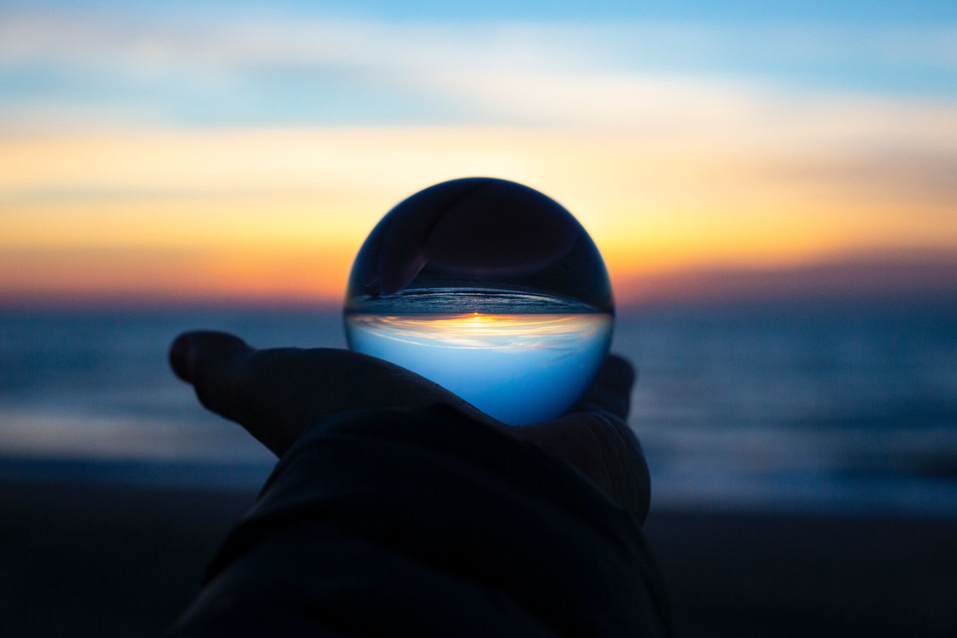 une main tenant une boule de cristal devant une mer, à l'arrière plan un lever de soleil 